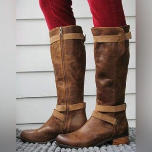 May include: Knee-high brown leather boots featuring a distressed finish, a side zipper closure, and two horizontal tan straps with buckle detailing. The boots have a low, stacked heel and a rounded toe, providing a structured look for the lower leg.