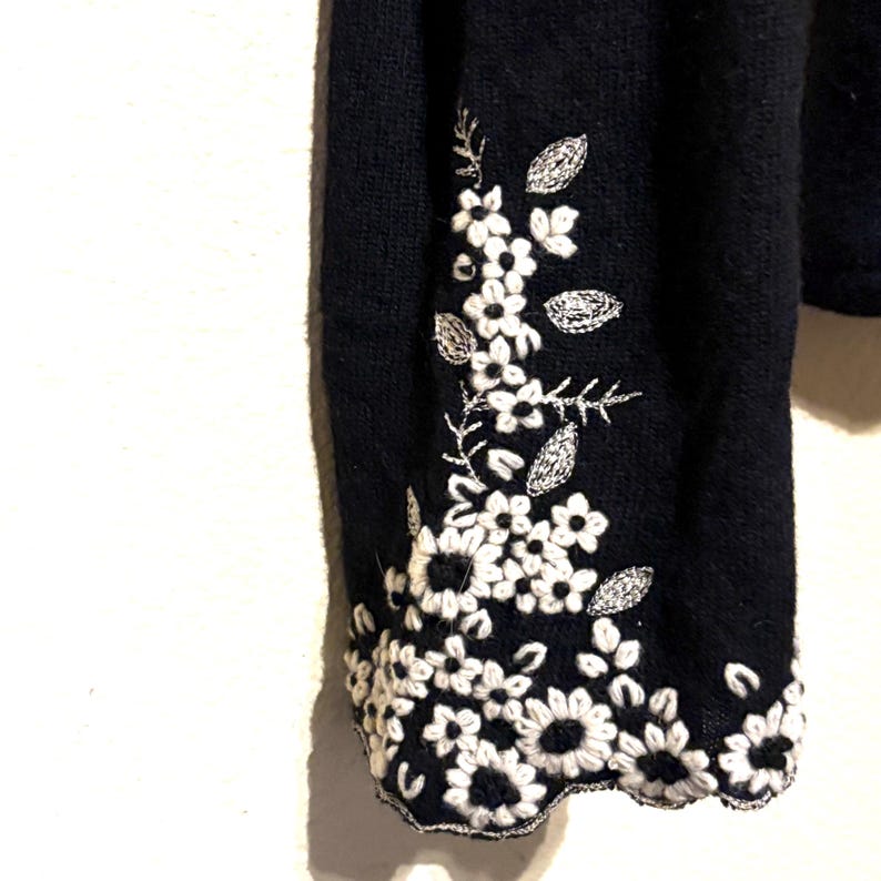 May include: Close-up of a black sweater sleeve with white floral embroidery. The embroidery features detailed flowers and leaves, creating a delicate pattern. The sleeve's edge has a scalloped design.