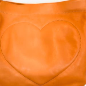 以下が含まれることがあります： オレンジ色の革製ショルダーバッグ。前面に大きなステッチのハートのデザイン。バッグは湾曲した上部とシルバーチェーンストラップを備えています。革は滑らかな質感で、暖かく鮮やかな色をしています。