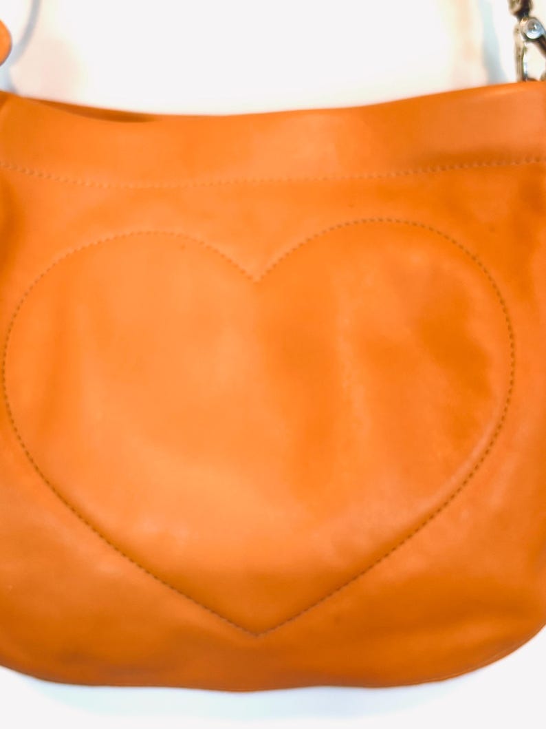 以下が含まれることがあります： オレンジ色の革製ショルダーバッグ。前面に大きなステッチのハートのデザイン。バッグは湾曲した上部とシルバーチェーンストラップを備えています。革は滑らかな質感で、暖かく鮮やかな色をしています。
