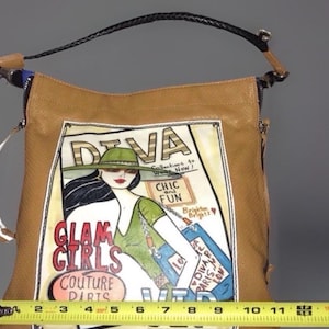 May include: A tan leather handbag with a black braided handle and a graphic print of a woman in a green hat. The print features the text "DIVA", "GLAM GIRLS", "COUTURE", "CHIC and FUN", and "Brighton". A yellow measuring tape shows a width of 11 inches.