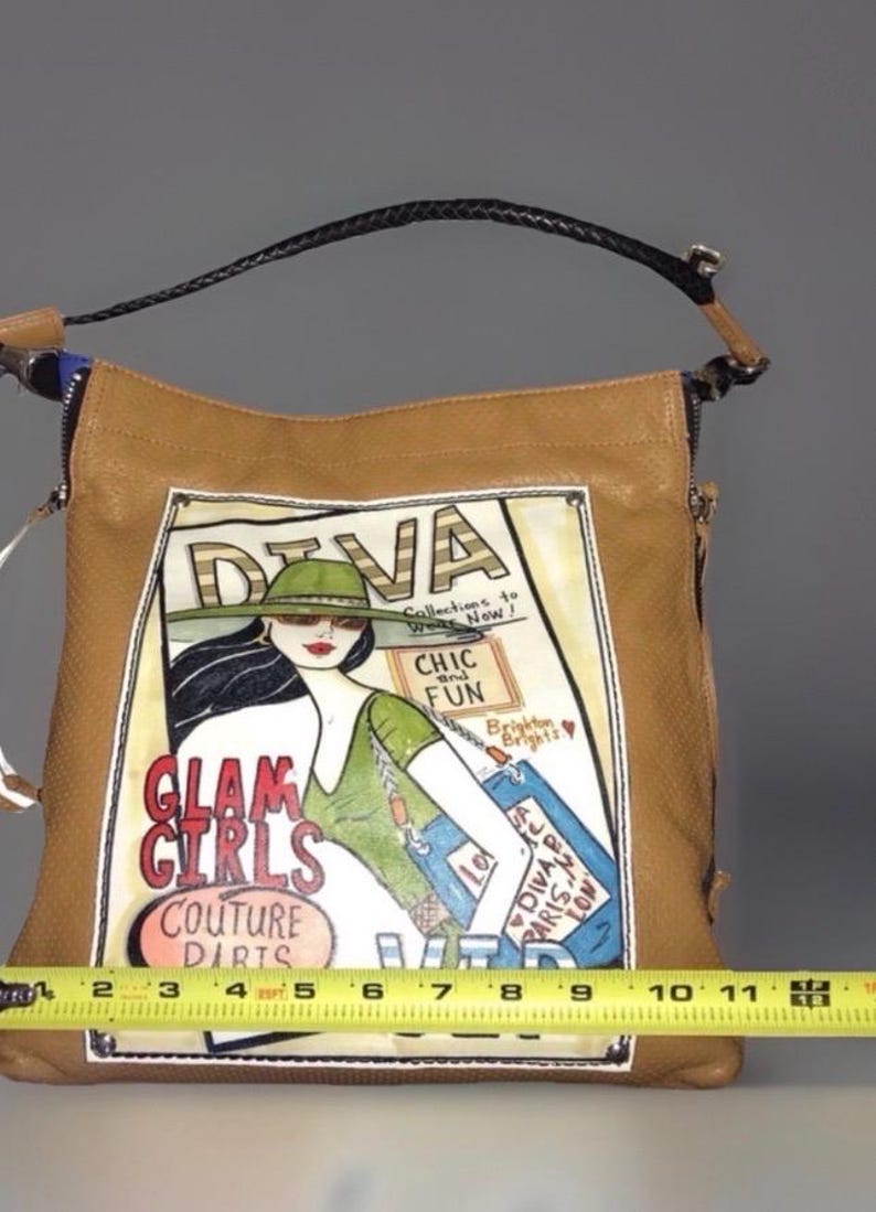 May include: A tan leather handbag with a black braided handle and a graphic print of a woman in a green hat. The print features the text "DIVA", "GLAM GIRLS", "COUTURE", "CHIC and FUN", and "Brighton". A yellow measuring tape shows a width of 11 inches.