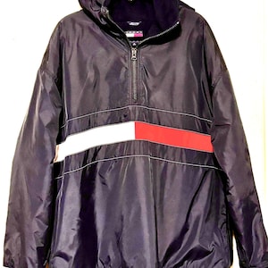 May include: Navy blue hooded jacket with a half-zip closure. The jacket features a white and red horizontal stripe across the chest. The cuffs have a small logo. The jacket is hanging on a gold hanger.
