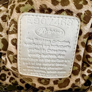 May include: A rectangular silver leather patch featuring the embossed text E907463 and Brighton, followed by a statement about the collection. The patch is set against a beige fabric lining with a brown and green leopard print pattern.