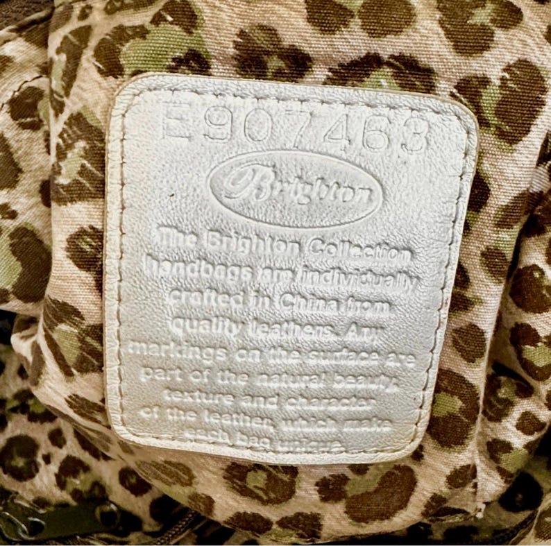 May include: A rectangular silver leather patch featuring the embossed text E907463 and Brighton, followed by a statement about the collection. The patch is set against a beige fabric lining with a brown and green leopard print pattern.