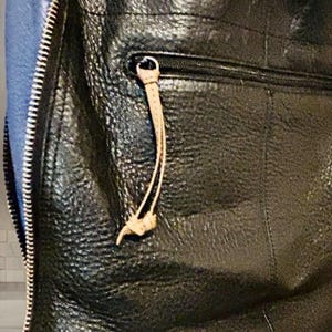 May include: A black textured leather bag featuring a vertical side zipper with a blue fabric panel. The front includes a horizontal zippered pocket with a light tan leather pull tab, showcasing a pebbled surface texture and visible stitching details.
