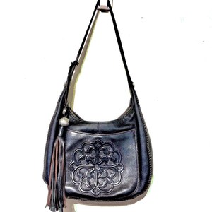 May include: Black leather shoulder bag with a decorative embossed floral design on the front. The bag has a long strap, a tassel, and a silver-toned charm. The bag is hanging against a white background.