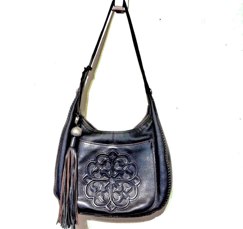 May include: Black leather shoulder bag with a decorative embossed floral design on the front. The bag has a long strap, a tassel, and a silver-toned charm. The bag is hanging against a white background.