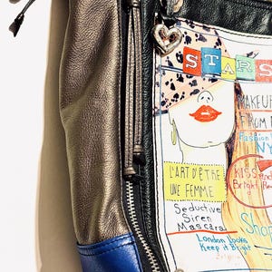 Pode incluir: Uma bolsa tote de couro multicolorida com fecho de z&iacute;per e um pingente em forma de cora&ccedil;&atilde;o. A bolsa apresenta uma ilustra&ccedil;&atilde;o de moda com a palavra "STARS" e o texto "MAKEUP FROM NY". A bolsa tem a parte inferior azul.