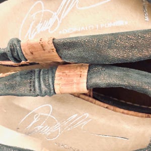 May include: Close-up of a pair of sandals with a dark teal upper and cork accents. The insole is beige with a white signature and the text "DONALD J PLINER". The sandals have a metallic sheen.