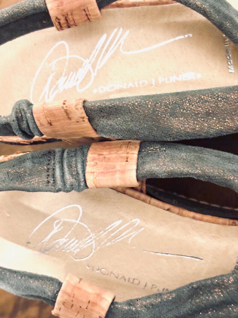 May include: Close-up of a pair of sandals with a dark teal upper and cork accents. The insole is beige with a white signature and the text "DONALD J PLINER". The sandals have a metallic sheen.
