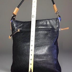 May include: A black leather shoulder bag with a textured surface, featuring a braided black strap with tan leather accents and silver-tone hardware. A yellow measuring tape is placed vertically over the bag, showing a height of 18 inches.