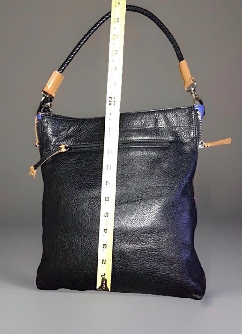 May include: A black leather shoulder bag with a textured surface, featuring a braided black strap with tan leather accents and silver-tone hardware. A yellow measuring tape is placed vertically over the bag, showing a height of 18 inches.