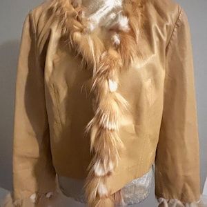 Könnte beinhalten: Hellbraune Jacke mit Fellbesatz am Kragen, vorne und an den Manschetten. Die Jacke scheint aus einem glatten Material zu bestehen, möglicherweise Leder oder einem lederähnlichen Stoff. Der Fellbesatz ist eine Mischung aus Hellbraun und Weiß.