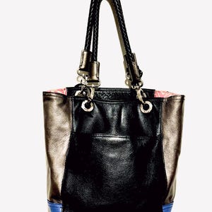 Pode incluir: Uma bolsa tote preta e met&aacute;lica com uma al&ccedil;a tran&ccedil;ada. A bolsa apresenta um painel central preto, pain&eacute;is laterais met&aacute;licos e um detalhe azul na parte inferior. O forro interno &eacute; rosa.