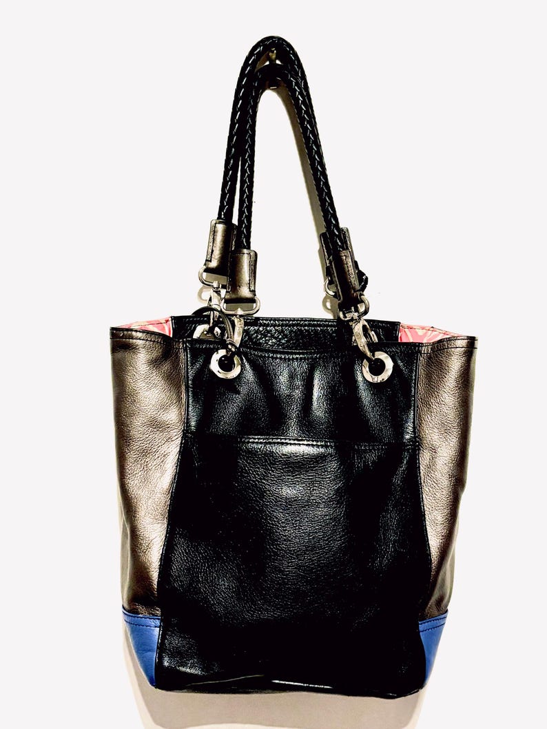 Pode incluir: Uma bolsa tote preta e met&aacute;lica com uma al&ccedil;a tran&ccedil;ada. A bolsa apresenta um painel central preto, pain&eacute;is laterais met&aacute;licos e um detalhe azul na parte inferior. O forro interno &eacute; rosa.