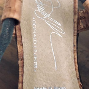 May include: Close-up of a sandal's insole with the brand name "DONALD J PLINER" and "Made in Spain" printed in silver. The insole is a light tan color, and the sandal has a cork and black strap design.