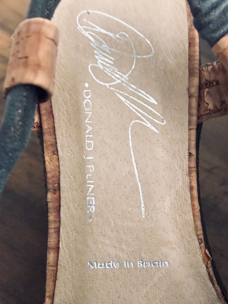 May include: Close-up of a sandal's insole with the brand name "DONALD J PLINER" and "Made in Spain" printed in silver. The insole is a light tan color, and the sandal has a cork and black strap design.