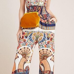 May include: A white jumpsuit with a colorful floral and peacock print. The jumpsuit has a fitted bodice and wide-leg pants. The model is wearing a yellow velvet clutch bag.