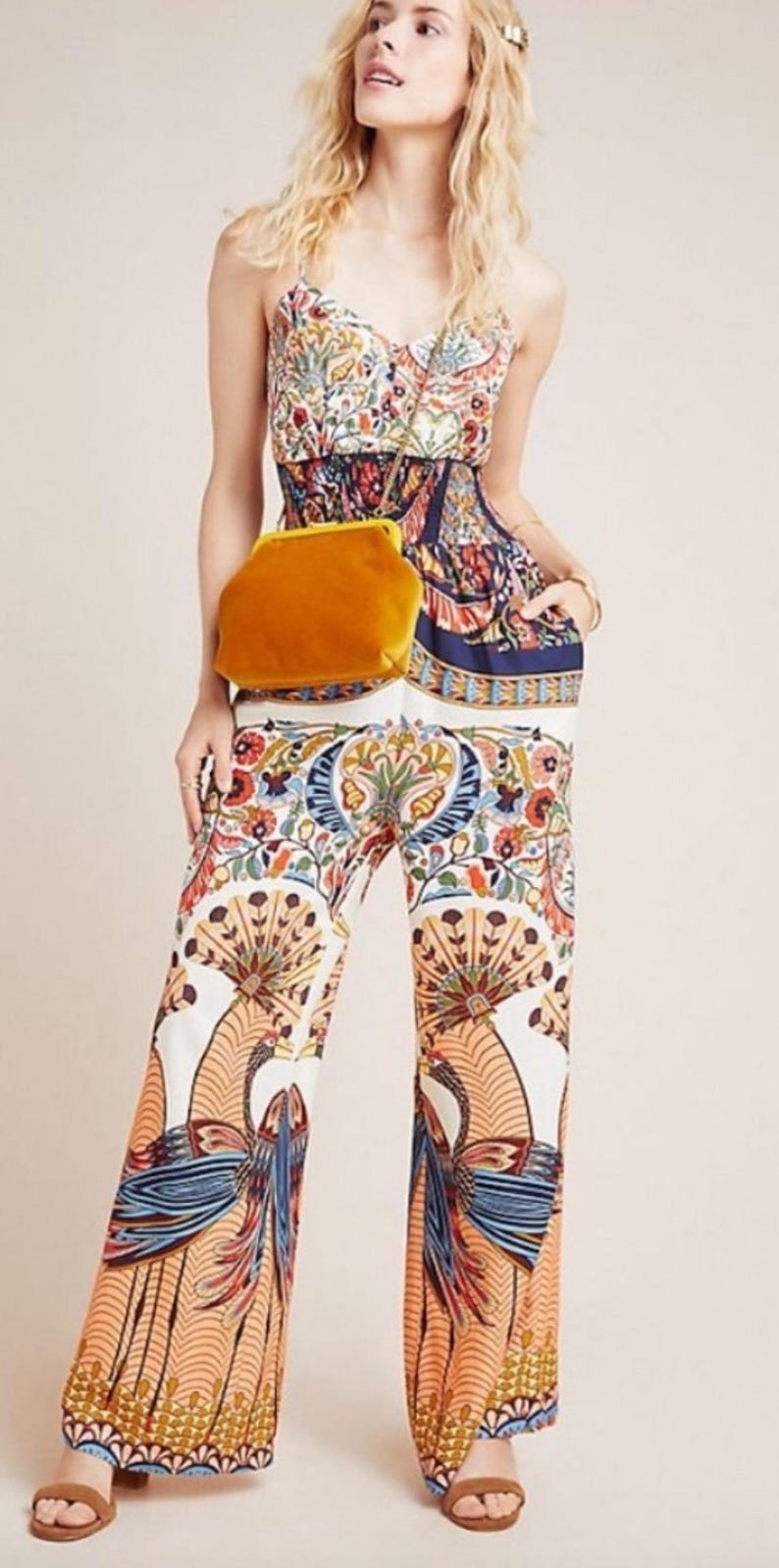May include: A white jumpsuit with a colorful floral and peacock print. The jumpsuit has a fitted bodice and wide-leg pants. The model is wearing a yellow velvet clutch bag.