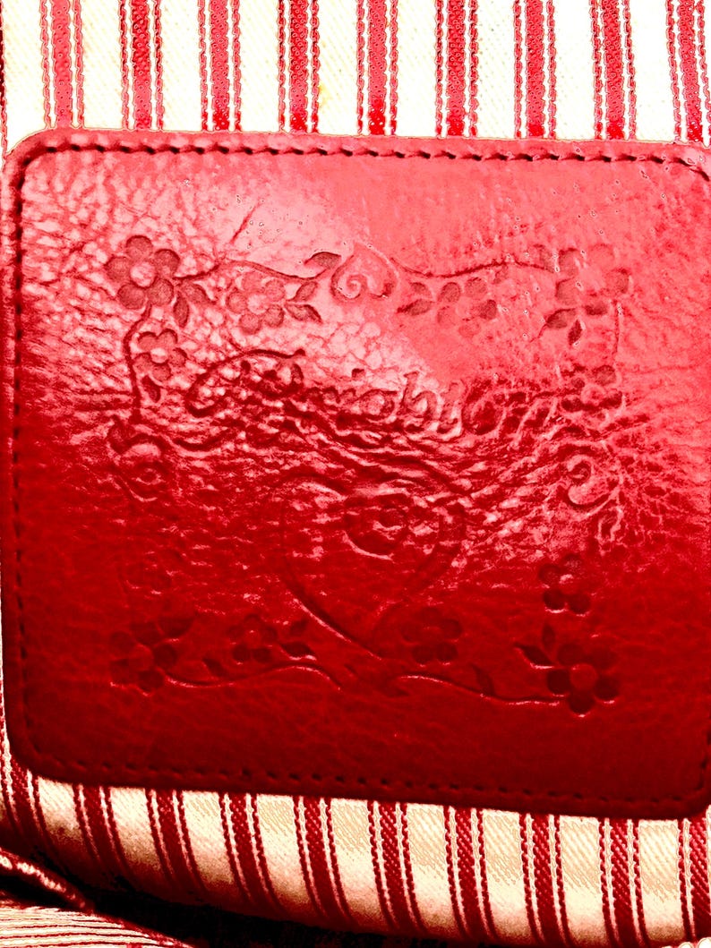 May include: A close-up of a red leather label with embossed floral designs and text. The label is stitched onto a red and white striped fabric. The textures and patterns create a vintage aesthetic. The overall look is handcrafted.