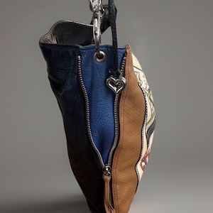 May include: A multi-colored leather handbag featuring panels of dark blue, bright blue, and tan perforated leather. The bag includes silver-tone metal zippers, a silver heart-shaped charm attached to a black strap, and a printed graphic design.