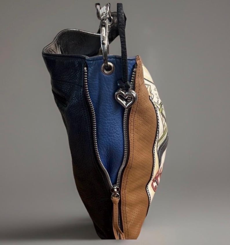 May include: A multi-colored leather handbag featuring panels of dark blue, bright blue, and tan perforated leather. The bag includes silver-tone metal zippers, a silver heart-shaped charm attached to a black strap, and a printed graphic design.