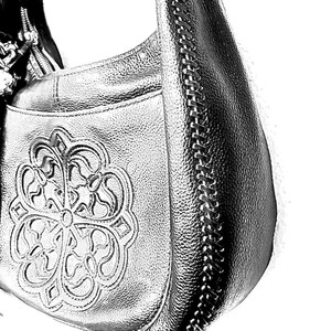 May include: Silver-colored leather hobo bag with a decorative floral design on the front. The bag features a chain detail along the side and a zipper closure. The bag has a textured surface.
