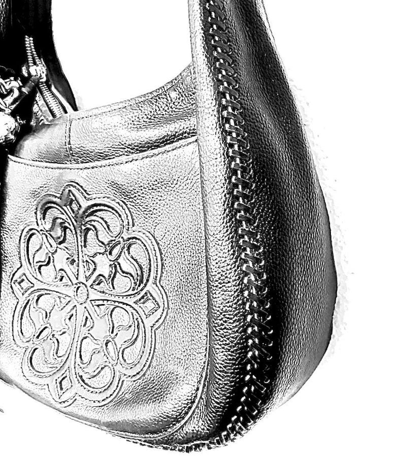 May include: Silver-colored leather hobo bag with a decorative floral design on the front. The bag features a chain detail along the side and a zipper closure. The bag has a textured surface.