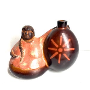 May include: A ceramic figurine depicting a woman holding a brown vessel. The woman has dark hair and is wearing a peach-colored garment. The vessel is brown with a sunburst design in orange. The figurine is on a white background.