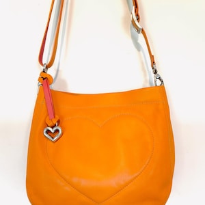 以下が含まれることがあります： 前面に大きなハートのデザインがステッチされたオレンジ色の革製ショルダーバッグ。バッグには、長い調節可能なストラップ、ピンクのアクセント、シルバーのハート型のチャームが付いています。バッグは鮮やかなオレンジ色です。