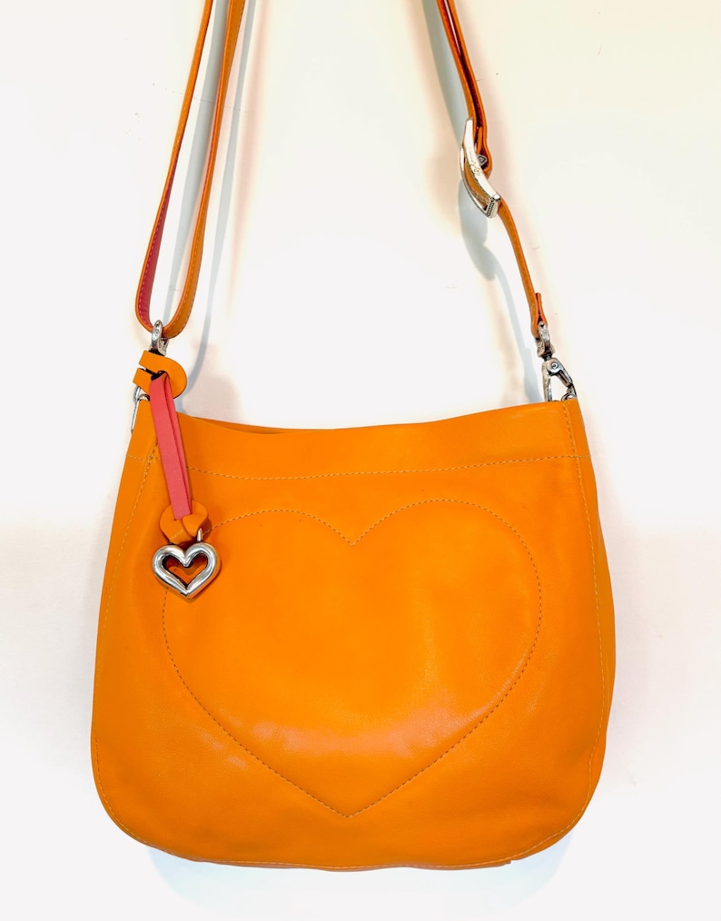以下が含まれることがあります： 前面に大きなハートのデザインがステッチされたオレンジ色の革製ショルダーバッグ。バッグには、長い調節可能なストラップ、ピンクのアクセント、シルバーのハート型のチャームが付いています。バッグは鮮やかなオレンジ色です。