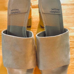 May include: A pair of light gray suede slip-on sandals with a woven heel. The brand name "jeffrey campbell" is printed on the insole. The sandals have a rounded toe and a thick heel.