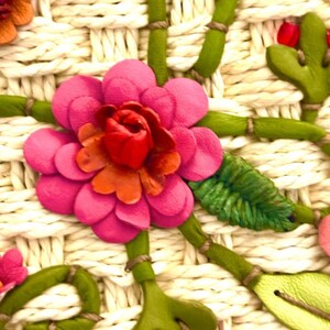 Puede incluir: Primer plano de una flor rosa con un centro rojo, tejida en un bolso de paja blanco. La flor est&aacute; rodeada de hojas y tallos verdes.