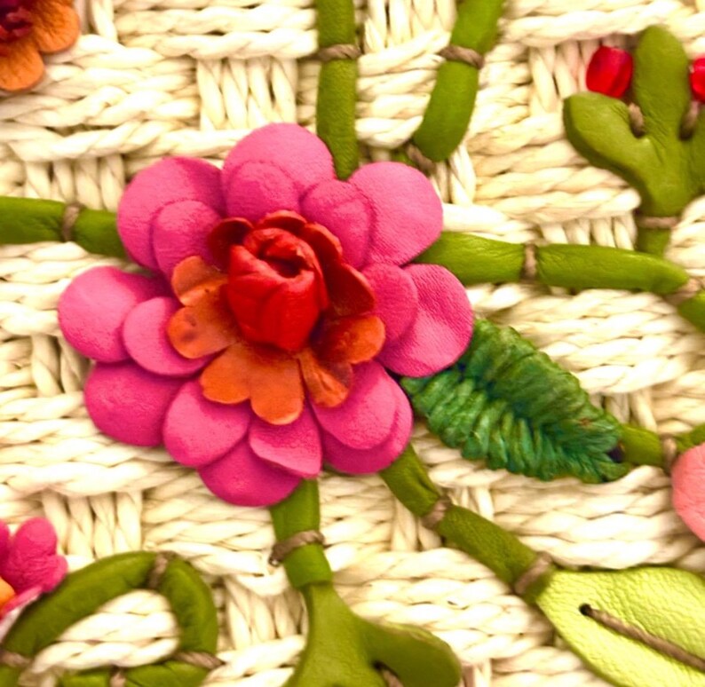 Puede incluir: Primer plano de una flor rosa con un centro rojo, tejida en un bolso de paja blanco. La flor est&aacute; rodeada de hojas y tallos verdes.