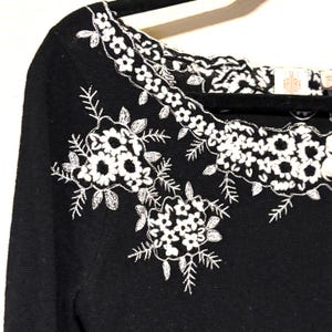 May include: Black sweater with white floral embroidery. The embroidery features detailed floral patterns around the neckline and on the front of the sweater. The sweater has a classic, elegant design.