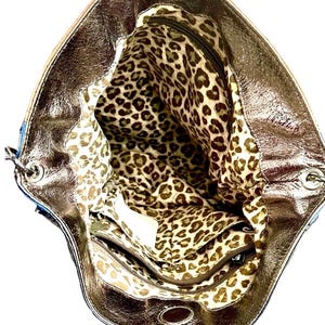May include: A metallic bronze handbag with a wide opening, revealing an interior lining featuring a brown and tan leopard print pattern. The bag includes a zippered interior pocket and silver-toned metal hardware at the strap attachment points.