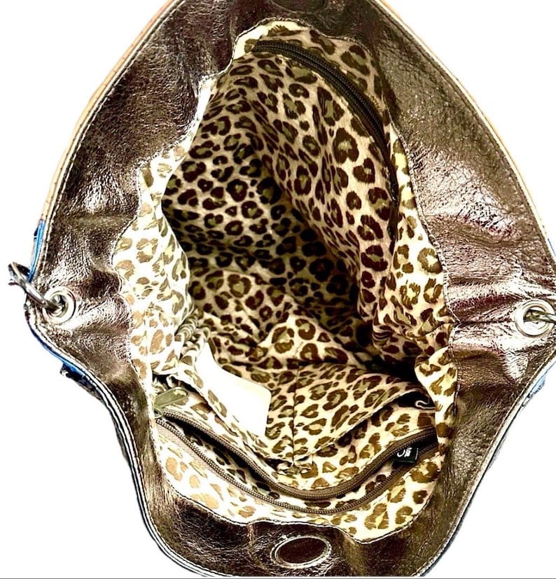 May include: A metallic bronze handbag with a wide opening, revealing an interior lining featuring a brown and tan leopard print pattern. The bag includes a zippered interior pocket and silver-toned metal hardware at the strap attachment points.