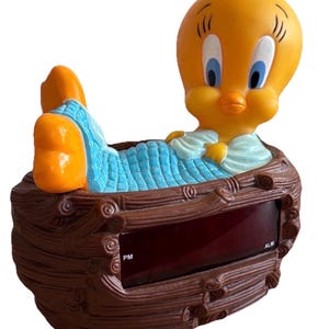 May include: A vintage alarm clock featuring the cartoon character Tweety Bird. The yellow bird is lying in a blue bed inside a brown, wooden-style frame. The clock face displays the time and has PM and ALM indicators.