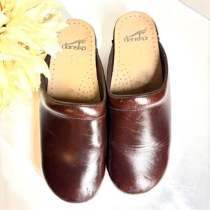 May include: A pair of brown leather clogs with the brand name "Dansko" visible on the insole. The clogs have a rounded toe and a smooth, glossy finish. The size is marked as 40. The background includes a soft, yellow floral arrangement.