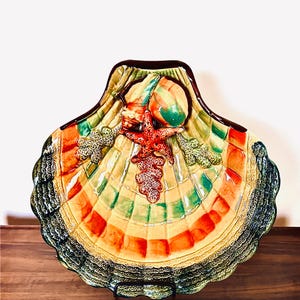 May include: A decorative seashell-shaped ceramic plate with a brown rim. The plate features a starfish, coral, and other sea life in shades of red, green, and orange. The plate is on a wooden surface.