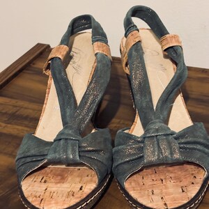 May include: A pair of teal-colored open-toe sandals with cork footbeds and a bow detail. The sandals have a slingback strap and a platform sole with white stitching. The upper straps are a darker teal with a metallic sheen.