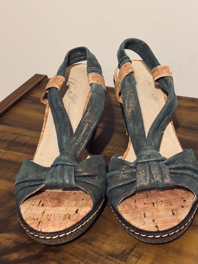 May include: A pair of teal-colored open-toe sandals with cork footbeds and a bow detail. The sandals have a slingback strap and a platform sole with white stitching. The upper straps are a darker teal with a metallic sheen.