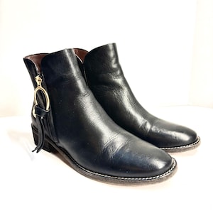 May include: A pair of black leather ankle boots. The boots have a side zipper with a gold-colored ring and tassel detail. The soles have white stitching. The boots are on a white background.