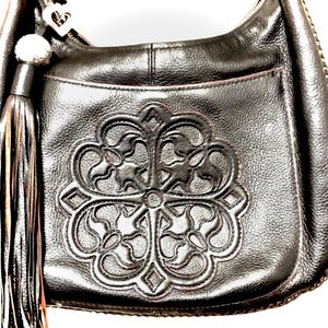 May include: Black leather shoulder bag with a decorative embossed floral design on the front. The bag features a long tassel and a silver-toned heart-shaped charm. The bag has a zipper closure and a pocket on the back.