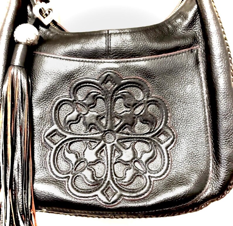May include: Black leather shoulder bag with a decorative embossed floral design on the front. The bag features a long tassel and a silver-toned heart-shaped charm. The bag has a zipper closure and a pocket on the back.