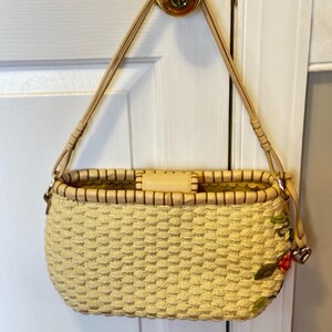 Puede incluir: Un bolso de mano de paja tejido con una correa de cuero marr&oacute;n claro y un detalle de bordado floral. El bolso tiene un cuerpo tejido de color crema y un ribete de cuero marr&oacute;n.