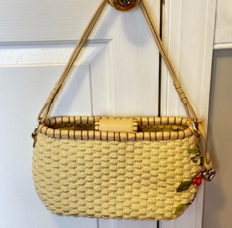 Puede incluir: Un bolso de mano de paja tejido con una correa de cuero marr&oacute;n claro y un detalle de bordado floral. El bolso tiene un cuerpo tejido de color crema y un ribete de cuero marr&oacute;n.