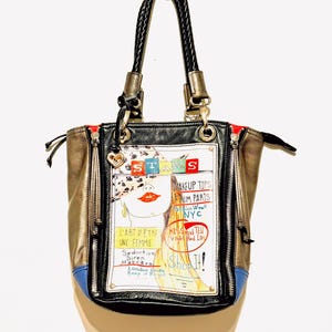 Pode incluir: Uma mala de m&atilde;o preta, dourada e azul com uma al&ccedil;a preta tran&ccedil;ada. A mala apresenta um gr&aacute;fico de uma mulher com o texto "STARS", "MAKEUP TIPS FROM PARIS NYC", "L'ART D'&Ecirc;TRE UNE FEMME" e "Shop It!"