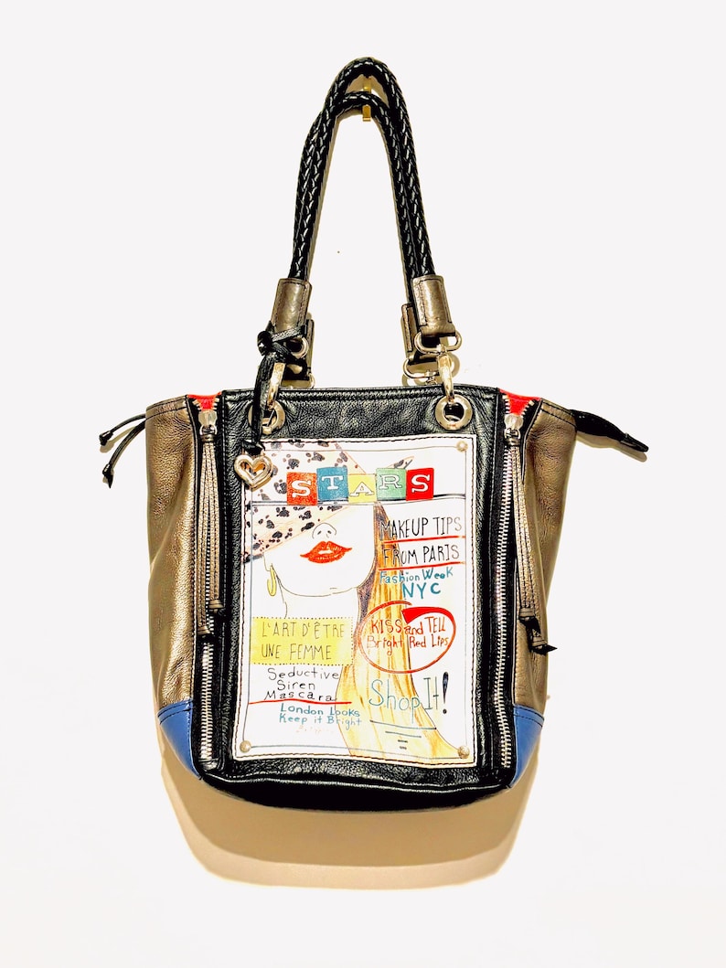 Pode incluir: Uma mala de m&atilde;o preta, dourada e azul com uma al&ccedil;a preta tran&ccedil;ada. A mala apresenta um gr&aacute;fico de uma mulher com o texto "STARS", "MAKEUP TIPS FROM PARIS NYC", "L'ART D'&Ecirc;TRE UNE FEMME" e "Shop It!"
